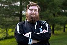 Justin Knapp—a Caucasian male with brown hair and a bushy beard—stands with his arms folded