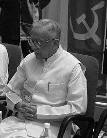 Greyscale image of an old Indian man, wearing a dhoti and kurta, and looking down.