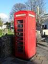 Three-quarter view of a red telephone booth with a glass door with red metal bars (partly in shadow) and a slightly domed roof. Behind it stand a flint wall, gravestones, a wooden bench and a tall stone war memorial.