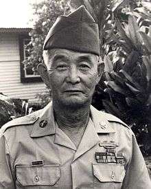 An older man in his military uniform with a hat on. There is a house and a tree in the background. He is looking forward at the camera.