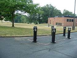 Color photograph of memorial (six posts with lights set around a rectangular demarcation) with grass, trees, and a building in the background.