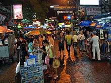 Night scene on a pedestrian street, with many people and street vendors; shops along the street bearing brightly lit signs with names like "99Fashion", "Brick Bar", "Mulligans Irish Bar", and Pepsi and McDonald's logos