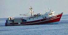 A small red fishing boat, without its usual tackle but with shipping containers at the rear, riding low astern in the open sea.