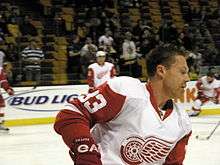 Hockey player in white and red uniform, with a picture of a puck with wings on his chest, without helmet. He skates across the ice, eyes closed.