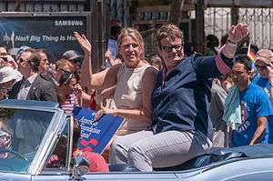 Two women sitting on the top of the back seat of an open-top automobile waving to a crowd.