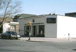 A bank building on the corner of a city street. A car can be seen parked out front and a traffic light is located on the sidewalk in front of the building. Other buildings can be seen in the background.