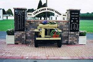 Front end of a jeep, sticking out of brick wall with plaques on brick pillars either side