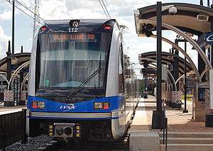 A blue and gray train stopped at a side platformed station with station black and gray station canopies visible.