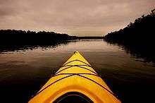 Triangular yellow prow of a boat in the foreground, on a smooth lake lined by dark trees under and reflecting a grey cloudy sky