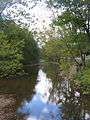 A smooth streem with some rocks in it flows between two wooded banks, reflecting the trees and clouds in the sky. A highway is at right.