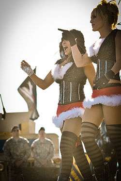 Two Caucasian women in matching outfits walk in front of two men in military uniforms. Both wearing black tops and red skirts with thigh-high black stockings, the woman on the left has dark brown hair and her hood up, while the blonde woman on the right is pointing to the left.