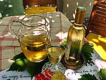 Colour photo illustrating well the song "Ah, the little white wine ...".  A carafe of white wine with a full glass and a corked bottle is placed on vine leaves, themselves on a white tablecloth with red tiles.  The bottle covered with mist gives a feeling of freshness and the play of light and shadow suggests that the scene is set under a Pergola.