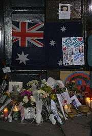 Dark doorway and doorstep with section of footpath. On the lower part of the dark door is a partly obscured Australian flag with dark blue background; red and white crosses on top left, large white star underneath and three white stars at the right with others obscured. Obscuring the right side of the flag is a white sheet with light blue writing, "Love + Miss You Always Heath" with two red hearts nearby, other writing includes "I'll never quit you" in darker blue, more writing is indistinct. This sheet also has six photos of a man. Above the flag, on the door is a smaller sheet with a photo of a man and indistinct writing below. On the doorstep and section of footpath are some 14 groups of flowers wrapped in plastic or paper, together with nine or so cards, five or six lit candles, and more photos. On the right side is a straw hat.