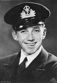 A head-and-shoulders portrait of a smiling man in naval uniform. He is wearing a cap that has a badge on the front depicting a crown that has an anchor hanging off it in the middle of splashing waves.