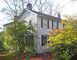 A white house with black shutters and brick chimneys seen from its front left corner. Shrubs and trees obscure the view on the sides.