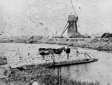 Black and white image of river showing small chain ferry on left bank with shire horse and driver ready to go across to the right bank where we can see a windmill