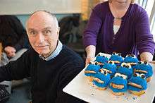 White male in his 70s, wearing a dark blue sweater, to the left of a woman holding a tray of Cookie Monster cupcakes