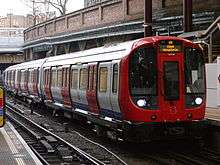 A train is slowing to stop at a platform on the right. Although there is a roof, sunlight can be seen through gaps; another platform and track can be seen on left. People are standing or walking on both platforms.