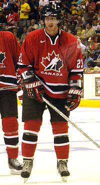 Hockey player in red Canada uniform. He stands on the ice, holding his stick on the ground, and looks to his right, lips slightly apart.