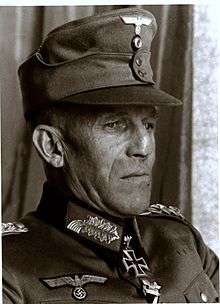 A grim looking man wearing a peaked cap and a military uniform with an Iron Cross displayed at his neck.