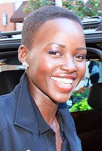 An African American female with a black automobile behind her is seen smiling. She is wearing a black coat.