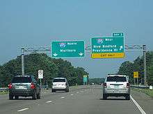 Ground-level view of three lanes of a divided freeway with two green exit signs directly overhead; the sign on the right is marked with a bright yellow banner that reads "Exit Only".