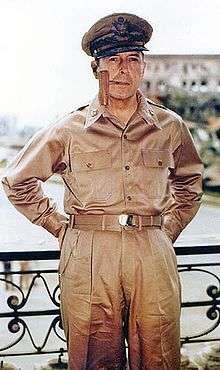 MacArthur in khaki trousers and open necked shirt with five-star-rank badges on the collar. He is wearing his field marshal's cap and smoking a corncob pipe.