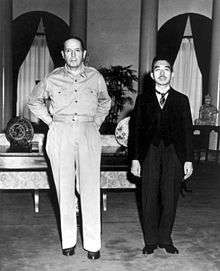 A tall Caucasian male (MacArthur), without hat and wearing open necked shirt and trousers, standing beside a much shorter Asian man (Hirohito) in a dark suit.