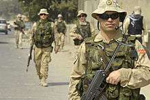 A five soldiers in tan colored desert camouflage and a green camouflage vests with dark sunglasses and tan hats patrol on a street.