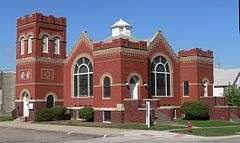 Red brick building with low square towers at two corners, somewhat taller square tower at a third, arched windows and doors