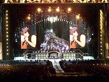 Distant shot of a concert stage. An arch with spotlights stands above a life-size locomotive and two big screens which display the male singer in left profile. Below and in front of the locomotive is musical equipment and four band members. At extreme left is a male playing guitar, he wears a school boy's uniform. Central stage has two guitarists with a drummer and his kit between them. Front stage has a catwalk with the singer turned to his left profile, he is cradling the microphone in his right hand and has his left fist near his face. He wears a cap, dark singlet and jeans.