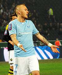 A photograph of a man wearing a sky blue sports shirt and white shorts on a football pitch.