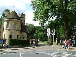 Turreted guardhouse at the open iron gate. People can be seen, walking with a background of trees.