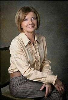 Polish First Lady - Maria Kaczyńska sitting on a chair wearing a light coloured, long sleeved top and long striped pants. The top has small ruffles on the cuffs and down the front. It has a shirt style collar. The chair is at an angle to the left and the subjects' head is turned toward the camera. Her legs are crossed and her arms are loosely crossed on her knees. She is smiling.