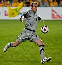 A man in a grey football kit in the act of kicking a ball.