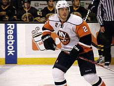 A Caucasian male ice hockey player shown from the knees up. He is wearing an orange, blue and white sweater with a white helmet.