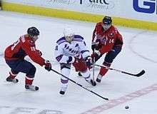 An ice hockey player, dressed in a white and blue jersey, stickhandles in between two opposing players, dressed in red jerseys. The defending players are on either side of the puck-handling player and have their sticks directed towards him.