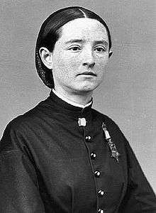 Head and shoulders of a white woman with straight hair pulled back into netting, wearing a tunic with a star-shaped medal hanging from a ribbon pinned to the left breast.