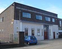 A brown-brick, flat-roofed, warehouse-style building. The lower half of its façade has been painted cream and decorated with arch-shaped window and door surrounds attached to the walls. On the first floor there are three two-pane windows and one single window. The side wall is mostly blank, with some small windows.