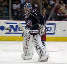 Hockey player in dark blue uniform and goaltender's gear. He stands on the ice with his hands on thighs; the left hand holds a hockey stick parallel to the rink.