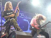 A color photograph of two members of the group Children of Bodom standing on a stage with guitars, drums are visible in the background