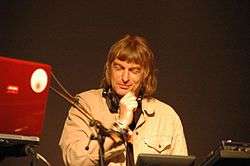 A brown-haired man with headphones around his neck sits in front of a mixer and a laptop.