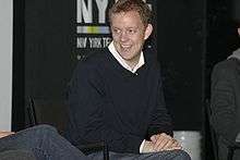 Matt Hubbard, a caucasian male with curly blond hair, wears a black sweater with a white shirt under. He smiles and looks to his right.