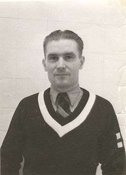 A photograph of a Caucasian man, wearing a sweater over a shirt and tie. He is pictured against a white tiled wall.