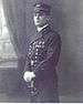 Side view of a white man standing with a sword in his hands, wearing white gloves, a cap, and a long military coat with a row of medals on the left breast.