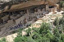 Ruins of circular and rectangular buildings under an overhanging cliff.