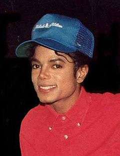 A man in a red shirt smiling toward the camera. Atop his head is a blue baseball cap.