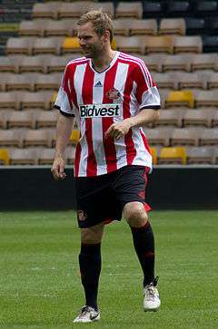 A man with light hair is wearing a red and white striped top, black shorts and black socks. He is standing on a grass field.