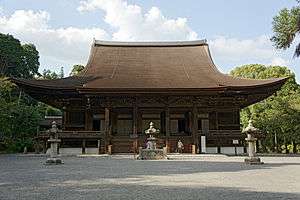 Wooden building with raised floor, a railed veranda and roofed stairs leading to the central entrance.