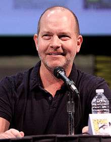 A man with closey shaven brown hair, speaking into a microphone, with his fist slightly raised above the table.